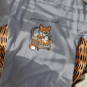 May include: Light blue t-shirt featuring a cartoon fox wearing glasses and reading a book while seated in a small armchair. The text "will process for snacks" is above the fox. The shirt is made of a soft material.