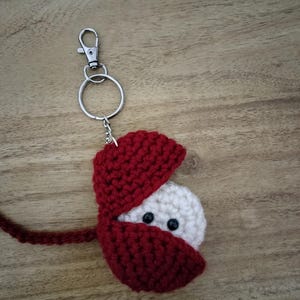 May include: A red crocheted heart-shaped keychain with a white crocheted face inside. The keychain has a silver metal ring and clasp.