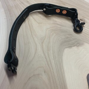 May include: A black leather camera strap with a metal buckle and two copper rivets. The strap has a metal clip on one end.