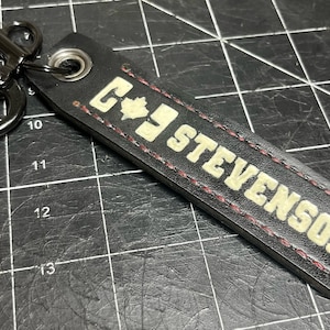 May include: Black leather keychain with the name "STEVENSON" in white block letters. The keychain has a metal clasp and a maple leaf design. The edges are stitched with red thread.
