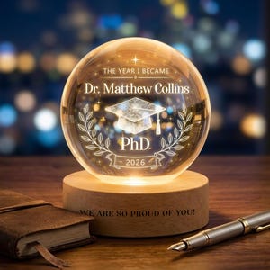 Könnte beinhalten: Ein personalisiertes Abschlussgeschenk. Eine klare Glaskugel mit dem Text "THE YEAR I BECAME Dr. Matthew Collins PhD 2026" und einem Abschlusskappen-Design. Die Kugel steht auf einem Holzsockel mit "WE ARE SO PROUD OF YOU!"