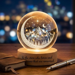 May include: A clear glass sphere lamp on a round wooden base featuring an etched mountain range, pine trees, a crescent moon, and stars. The base is engraved with the text: To the stars who listen and dreams that are answered.