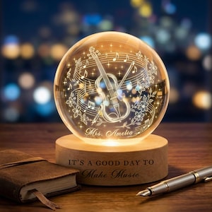 May include: A clear glass sphere featuring an internal laser-etched violin, musical notes, and floral patterns, with the name Mrs. Amelia engraved. The sphere rests on a light wood base inscribed with the text IT'S A GOOD DAY TO MAKE MUSIC.
