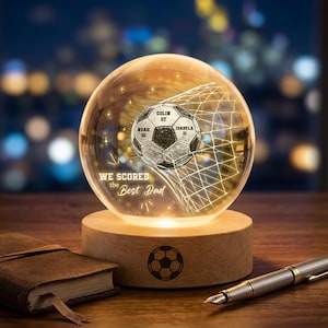 Può includere: Una sfera di vetro trasparente con un design a pallone da calcio, personalizzata con nomi e numeri, poggia su una base di legno. La base ha una grafica a pallone da calcio. Un taccuino rilegato in pelle e una penna sono in primo piano.