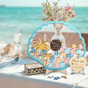 May include: A wooden seashell-shaped guest book with a blue border, featuring palm trees and star cutouts. The text "Tim and Courtney July 15, 2026" is displayed on a circular tag. A small wooden box and a heart-shaped sign are also present.