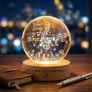 Puede incluir: Una esfera de cristal transparente con notas musicales y un diseño de clave de sol, apoyada sobre una base de madera. La base tiene el texto "It's a good day to make music". Un bolígrafo y un cuaderno de cuero están sobre la mesa.