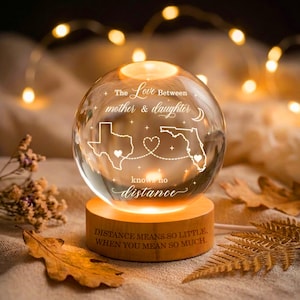 Op de afbeelding: Een heldere glazen bol rustend op een cilindrische houten voet. De bol is gegraveerd met de tekst The Love Between mother & daughter knows no distance, kaarten van Texas en Florida, en een hartje. De voet bevat de tekst DISTANCE MEANS SO LITTLE, WHEN YOU MEAN SO MUCH.