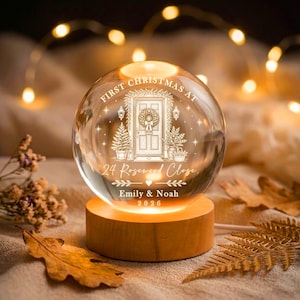May include: A clear glass sphere ornament on a round wooden base, featuring an engraved illustration of a front door with a wreath, flanked by potted trees. Text reads: FIRST CHRISTMAS AT 24 Rosewood Close, Emily & Noah, 2026, with a small heart.