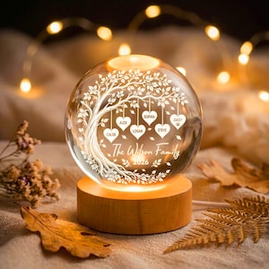 Könnte beinhalten: Ein kugelförmiges Glas-Nachtlicht auf einem zylindrischen Holzsockel. Das Glas zeigt ein graviertes Baummotiv mit hängenden herzförmigen Ornamenten, beschriftet mit Caleb, Alex, Logan, Sophia, Steve und Daisy, sowie dem Text The Wilson Family 2026.
