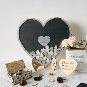 May include: A wooden heart-shaped guest book with a gray background and a silver frame. The heart is filled with small white and gray wooden hearts. The sign reads "Please sign our guest book".