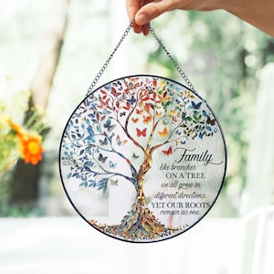 Könnte beinhalten: Runde, dekorative Glasscheibe mit einem Baumdesign mit bunten Blättern und Schmetterlingen. Der Baum hat einen braunen Stamm und Wurzeln, mit dem Text "Family like branches on a tree..." in einer Schreibschrift. Eine schwarze Kette ist zum Aufhängen befestigt.