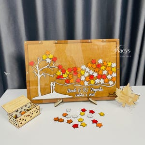 May include: Wooden wedding guest book with a tree and leaf design. The book features a clear acrylic front and includes a box for leaf-shaped guest signatures. The names "Nicole & RJ Fagerlie" and the date "October 4, 2025" are displayed.