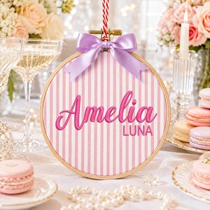 Pode incluir: Um bastidor de bordado redondo de madeira contendo tecido com listras verticais rosa e brancas. O nome Amelia está bordado em letra cursiva rosa escuro, com o nome LUNA bordado abaixo em letras de forma. Um laço de cetim lilás está preso.