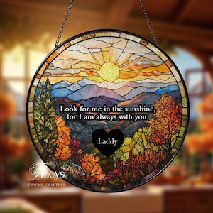 Puede incluir: Una ventana redonda de vidrieras con marco y cadena negros. La obra representa una escena de puesta de sol con montañas, árboles y el texto "Look for me in the sunshine, for I am always with you." Un corazón negro con el nombre "Laddy" está en el centro.