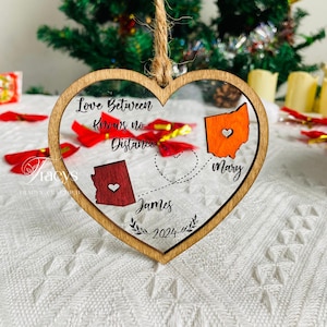 May include: Heart-shaped wooden ornament with a clear center, featuring the text "Love Between Knows no Distance." It includes state outlines with hearts, names, and the year 2024. A twine loop is attached for hanging.