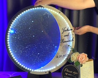 May include: A round, illuminated wedding sign with a crescent moon design. The sign features the names "Jackson and Savannah" and the date "9.21.2025". The sign is supported by a black stand and surrounded by decorative elements.