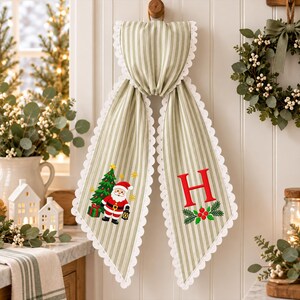May include: A sage green and white striped fabric hanging decoration with white scalloped edges. One side features an embroidered Santa Claus with a Christmas tree, while the other side displays a large red letter H above a sprig of holly berries.