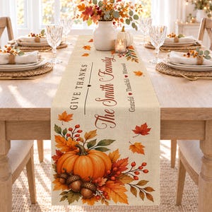 Könnte beinhalten: Ein beigefarbener Tischläufer aus Leinen mit herbstlichem Aufdruck, bestehend aus einem großen orangefarbenen Kürbis, Eicheln und Herbstblättern. Der Text lautet Give Thanks, The Smith Family und Grateful Thankful Blessed, ergänzt durch rote Herzen.