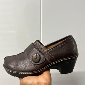 May include: A dark brown leather clog with a textured surface, featuring a wide strap across the instep secured by a single round button. The shoe has a thick, dark sole with visible white contrast stitching along the base and a block heel.