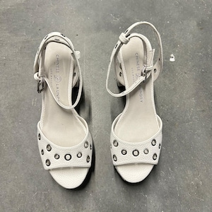 May include: A pair of white textured block heel sandals featuring silver-tone metal grommets across the toe strap and ankle strap. The insole is branded with the text Chinese Laundry Los Angeles. The sandals have an adjustable ankle strap buckle.