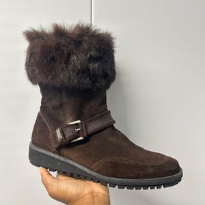May include: A dark brown suede mid-calf boot featuring a thick, fluffy dark brown fur cuff around the top opening. The boot includes a decorative side strap with a silver-toned metal buckle and a textured black rubber sole with a treaded bottom.