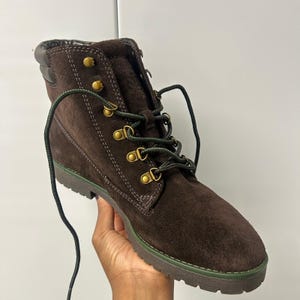May include: A dark brown suede ankle boot featuring a padded collar, brass-toned metal eyelets, and dark green laces. The boot has a thick, lugged brown sole with a thin green accent stripe running along the side of the midsole construction.