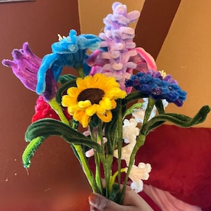 Puede incluir: Un colorido ramo de flores hechas a mano con limpiapipas. El arreglo incluye un girasol amarillo, iris azules, tulipanes morados y flores blancas, con tallos verdes. Las flores se sostienen en una mano sobre un fondo rojo y marrón.