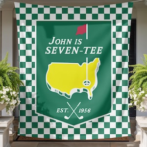 May include: A rectangular banner featuring a green and white checkered border surrounding a central green shield. The shield displays a yellow map of the United States, a red flag, two crossed golf clubs, and the text John is SEVEN-TEE EST. 1956.