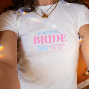 Puede incluir: Una camiseta blanca de manga corta con el texto BOMBSHELL BRIDE en letras rosas y azules, junto con el texto Katy HAS ENTERED THE VILLA impreso debajo en una combinación de fuentes azules y rosas en el área del pecho de la prenda.
