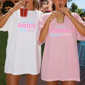 May include: A white t-shirt with the text BOMBSHELL BRIDE Katy has entered the villa in blue and pink, and a pink t-shirt with the text HOT NEW BOMBSHELL Hannah has entered the villa in blue and pink. Both shirts have short sleeves and crew necks.
