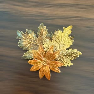 May include: A metallic brooch featuring two textured maple leaves in shades of gold and bronze, layered behind a central multi-petaled flower with a detailed stamen center. The piece displays a polished finish with varying metallic tones throughout.