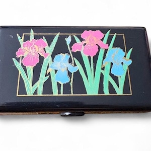 May include: A rectangular black case featuring a gold-outlined rectangular frame containing a floral illustration of two pink irises and two blue irises with green stems and leaves. The case has a smooth, dark finish and a small central latch.