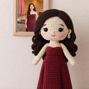 May include: A handcrafted crochet doll with dark brown curly hair and a burgundy dress. The doll has a cream-coloured face with rosy cheeks and is wearing silver earrings. A framed photo of a woman in a burgundy dress hangs in the background.