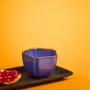 May include: A small, hexagonal, blue ceramic bowl with a gold rim sits on a dark rectangular tray. A piece of red pomegranate is on the tray. The background is a solid orange color.