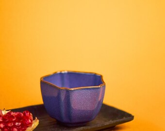 May include: A small, hexagonal, blue ceramic bowl with a gold rim sits on a dark rectangular tray. A piece of red pomegranate is on the tray. The background is a solid orange color.