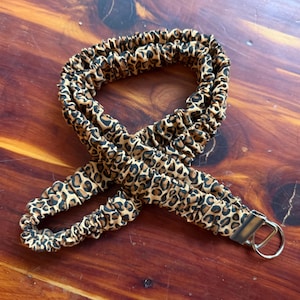 May include: A leopard print strap with a metal clasp and ring. The strap is tan with brown and black spots. The strap is made of a stretchy material.