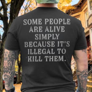 Könnte beinhalten: Ein schwarzes Kurzarm-T-Shirt mit dem Text in verwaschener grauer Schrift: SOME PEOPLE ARE ALIVE SIMPLY BECAUSE IT'S ILLEGAL TO KILL THEM. Das Shirt wird von der Rückseite gezeigt, wobei die Botschaft deutlich in der Mitte zu sehen ist.