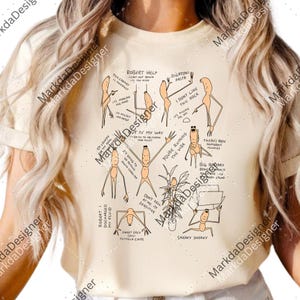 May include: Cream-colored t-shirt with a humorous cartoon design. The graphic features several stick figures with various expressions and speech bubbles, including phrases like "Robert Help" and "Sneaky Sneaky."