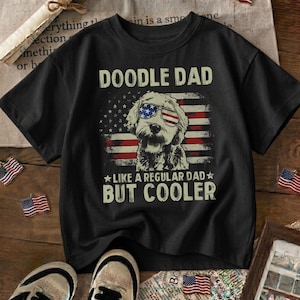 May include: Black t-shirt with the text "DOODLE DAD" above an illustration of a dog wearing American flag sunglasses. The dog is in front of an American flag design. The text "LIKE A REGULAR DAD BUT COOLER" is below the dog.