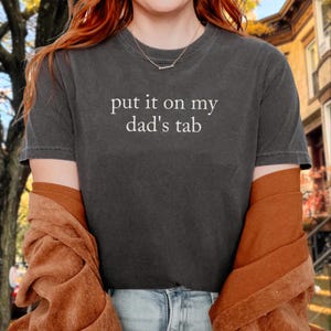 Puede incluir: Camiseta gris oscuro con el texto blanco "put it on my dad's tab". La camiseta tiene mangas cortas y cuello redondo. Una chaqueta marrón está sobre los brazos.