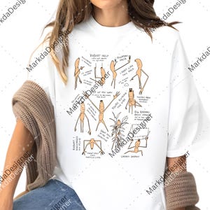 May include: White t-shirt featuring a humorous cartoon design with stick figures and various phrases. The design includes text like "ROBERT HELP" and "I DON'T LIKE THIS ROCK". The shirt is a casual, everyday wear item.