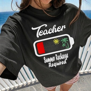 Puede incluir: Camiseta negra con la palabra "Teacher" en escritura blanca sobre un gráfico de batería. La batería muestra una carga baja con un sol y palmeras, y el texto "Summer Recharge Required" debajo.