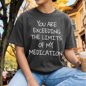 Może przedstawiać: Ciemnoszary t-shirt z krótkim rękawem z białym, odręcznym napisem: "YOU ARE EXCEEDING THE LIMITS OF MY MEDICATION". Koszulka ma okrągły dekolt i luźny krój, wykonana jest z miękkiej, jednokolorowej tkaniny bawełnianej o gładkiej fakturze.