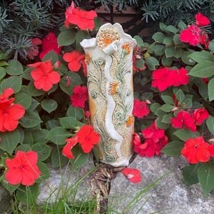 May include: A tall, cylindrical ceramic vase featuring a cream-colored base with intricate, raised relief patterns of green vines, swirling stems, and orange floral accents. The surface displays textured detailing and a glossy finish.