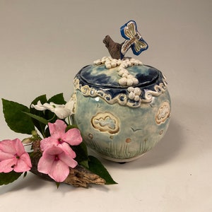 May include: A ceramic jar with a lid decorated with a blue dragonfly, white clouds, and a blue and white sky. The jar is sitting on a white surface with a green plant and pink flowers.