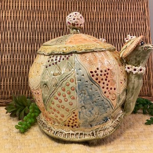 May include: A ceramic teapot featuring a patchwork design with various textures, including dots, lines, and floral patterns in shades of peach, green, and blue. The lid has a small floral knob, and the spout is shaped like a turtle wearing a hat.