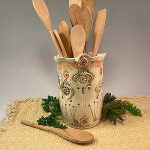May include: A cylindrical ceramic utensil holder with a cream-colored glaze and intricate embossed patterns in orange and green. The holder is filled with various light-colored wooden spoons and spatulas, with one additional wooden spoon lying flat.