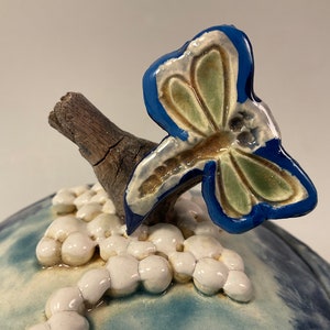 Pode incluir: Um pote de cerâmica com tampa com um esmalte azul e branco. A tampa apresenta uma libélula azul e verde pousada em um galho.