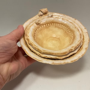 Może przedstawiać: Zestaw trzech ceramicznych misek w ciepłym, beżowym odcieniu. Miski mają teksturowany, organiczny design z falistą krawędzią. Najmniejsza miska ma dekoracyjne obramowanie. Miski są ułożone w stos.