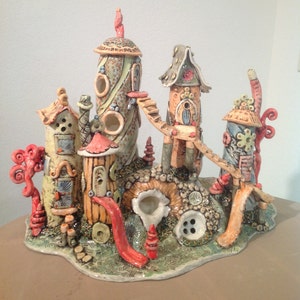 May include: A whimsical ceramic sculpture of a miniature village with colorful houses, a slide, and a bridge. The houses are decorated with buttons, beads, and other details. The sculpture is set on a green base with a textured surface.